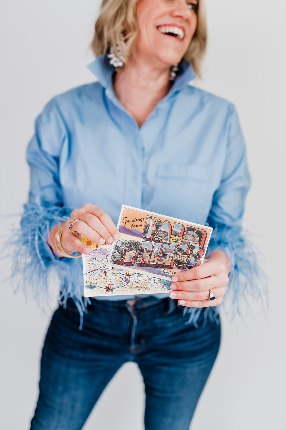 a woman smiling looking away from the camera holding up palm springs post cards