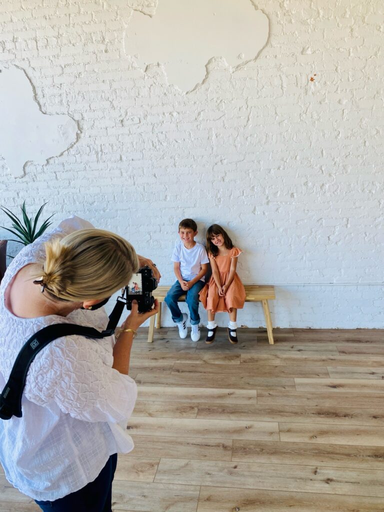 attending-a-photography-retreat Photographer captures two smiling children sitting on a bench against a textured white brick wall backdrop while attending a photography retreat
