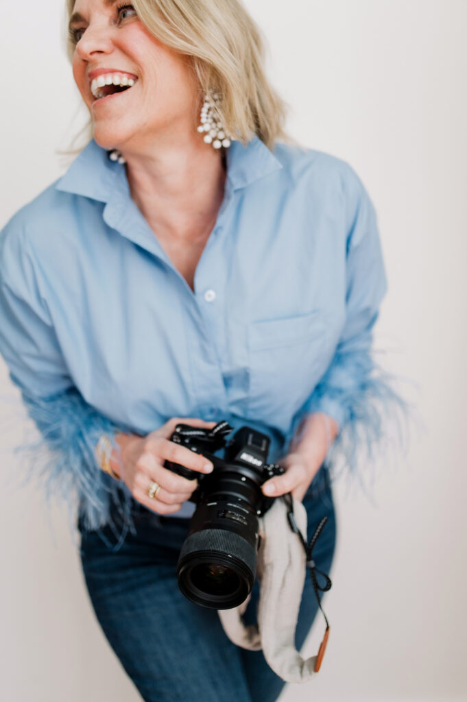 Introducing…Four Must-Have Website Resources to Uplevel Your Client Experience - NPS Photography Laughing photographer in a feather-trimmed blue shirt holding a camera, embodying trusted photography website resources.