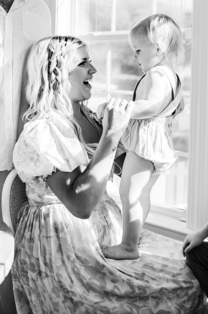 Summer-family-photography-in-DC A joyful black-and-white moment of a mother laughing with her toddler by a sunlit window.