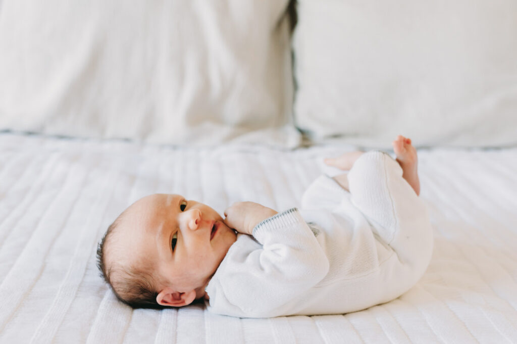 in-home-photo-shoot A newborn lies peacefully on a soft white blanket, gazing curiously at the camera in natural light.