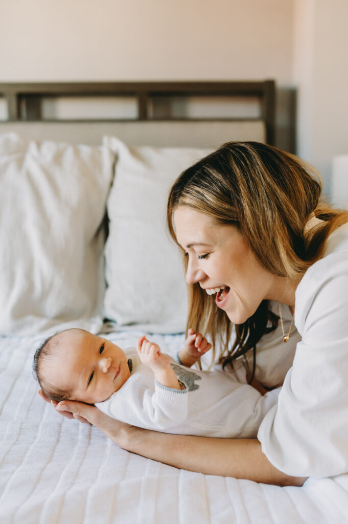 in-home-photo-shoot A joyful mother engages with her newborn during a warm and natural in-home photo shoot on the bed.
