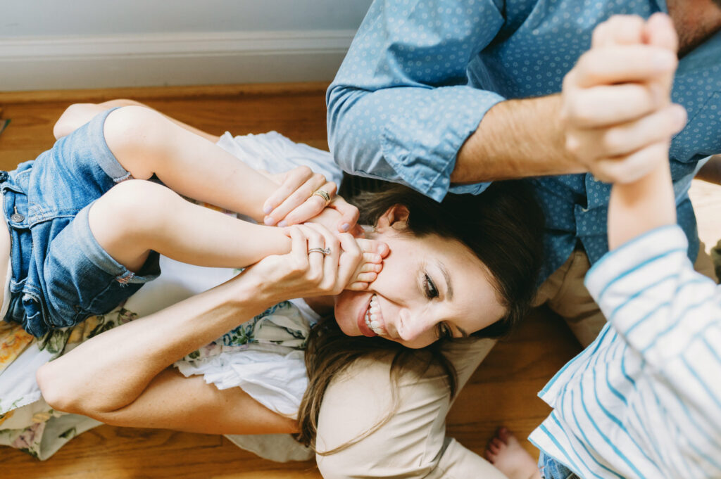photography-prompts-for-kids A playful mom laughs while holding her child’s foot near her face during a joyful family moment at home.