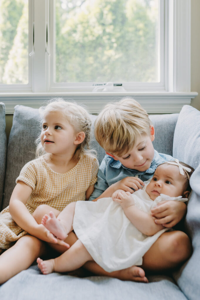 What to Expect After Your Photo Session: A Step-by-Step Guide - NPS Photography Older siblings gently hold their newborn sister on the couch, illustrating what to expect after your photo session with candid, loving moments.
