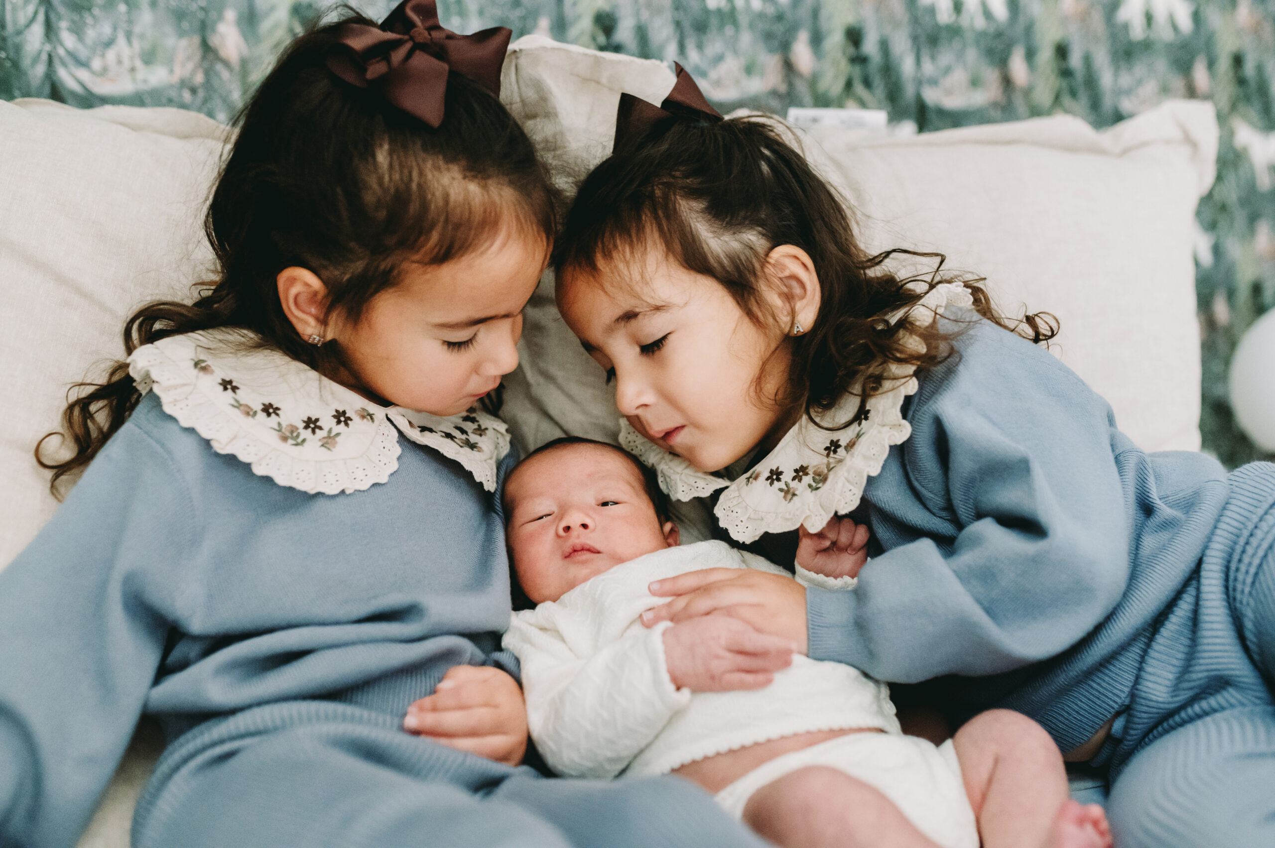 Two big sisters in matching outfits snuggling their newborn sibling during an in-home session—a reminder of why knowing when to book a newborn photographer ensures these first sibling moments are never missed.