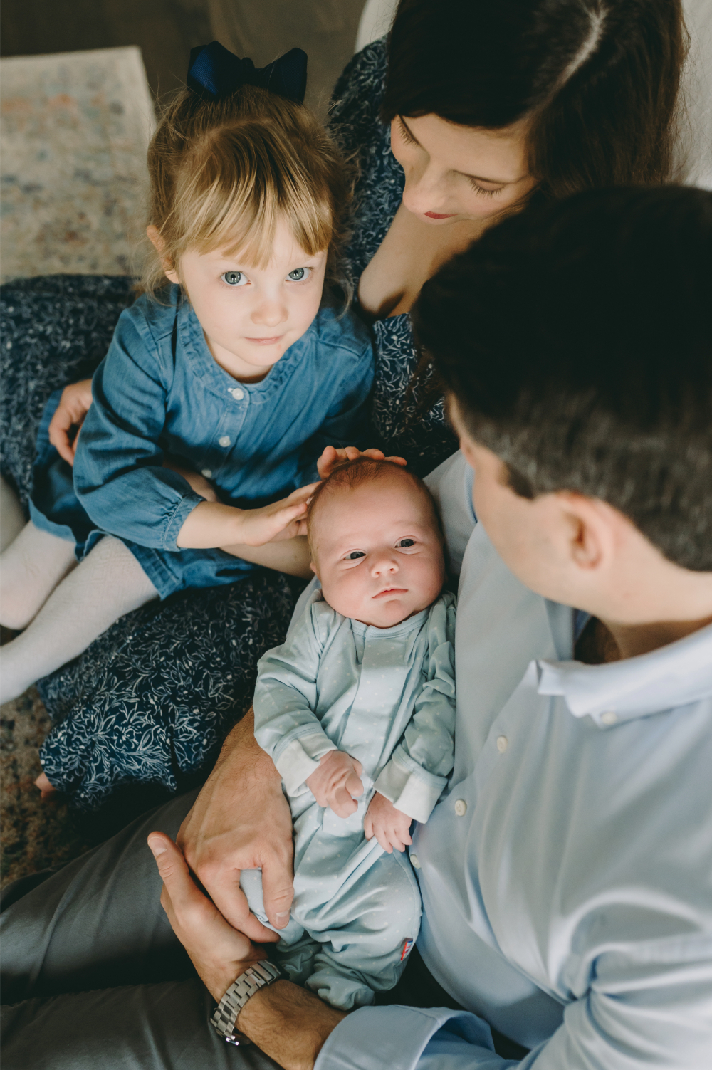 A toddler gently touches her newborn brother’s head as parents look on with love—an intimate family moment captured because they knew when to book a newborn photographer in advance.