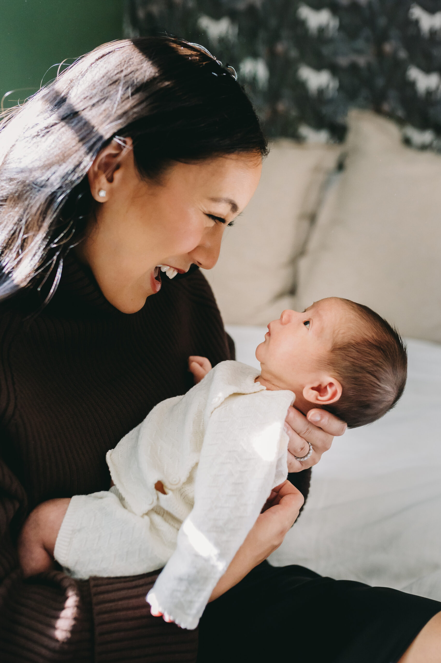 A joyful mom gazes at her newborn in natural light during an in-home session, showing how special moments are possible when families know when to book a newborn photographer.