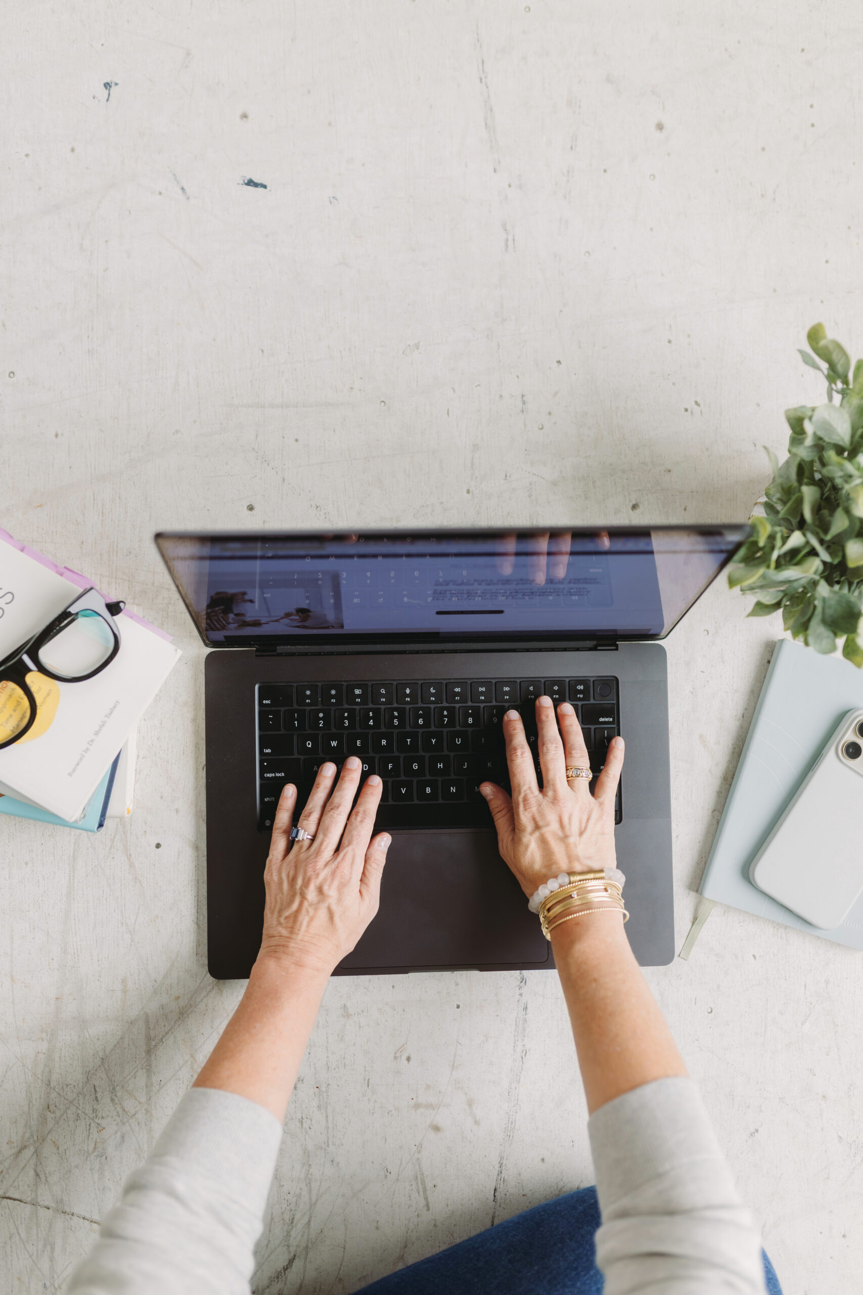 What’s the Best CRM for Photographers? Here’s My 2025 Review - NPS Photography Hands typing on a MacBook surrounded by glasses, notebooks, and a phone—illustrating behind-the-scenes work with the best CRM for photographers.