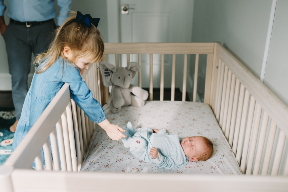 Why Full-Service Family Photography Is Worth Every Penny (And Then Some) - NPS Photography Big sister gently checks on her newborn sibling in the crib during a relaxed in-home session.