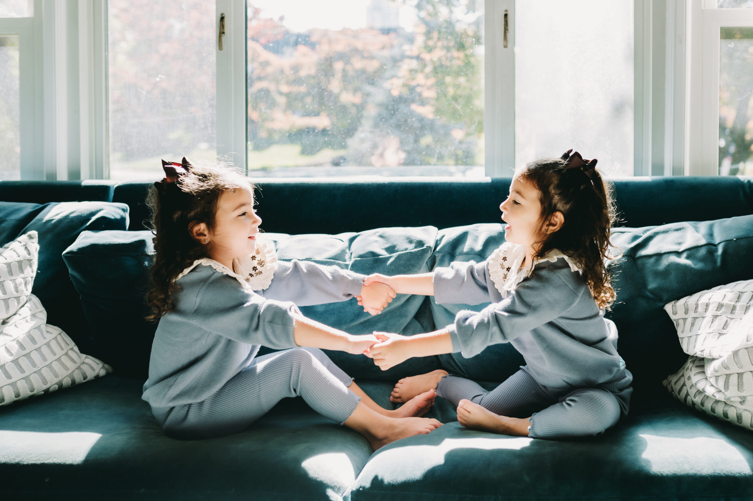 Toddler twins play and giggle on a velvet couch in the sunlight during a relaxed full-service family photography session.
