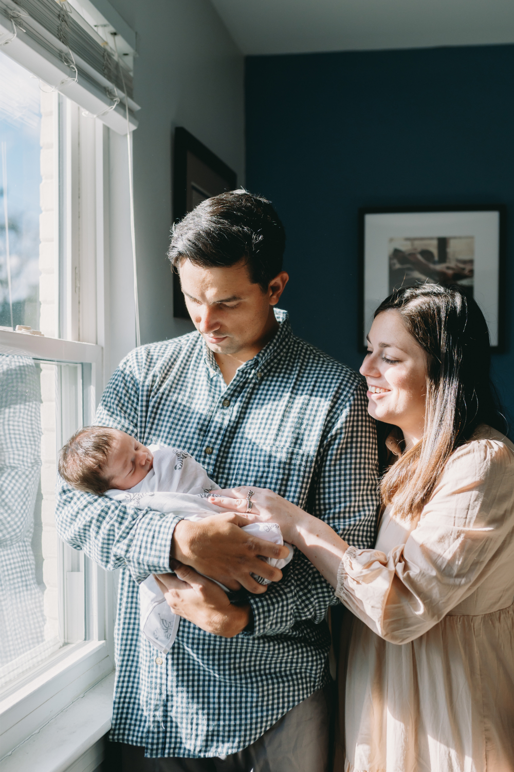 Why Full-Service Family Photography Is Worth Every Penny (And Then Some) - NPS Photography New parents cradle their newborn by the window during a natural light full-service family photography session.