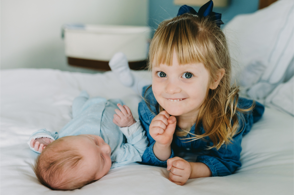 Big sister smiles next to her newborn sibling during cozy in-home photography client experiences.