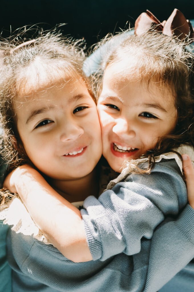 Photography Ideas In Home: Helping Shy Kids Shine During Family Sessions - NPS Photography Twin girls hugging tightly with big smiles in natural light—capturing sibling love through photography ideas in home.