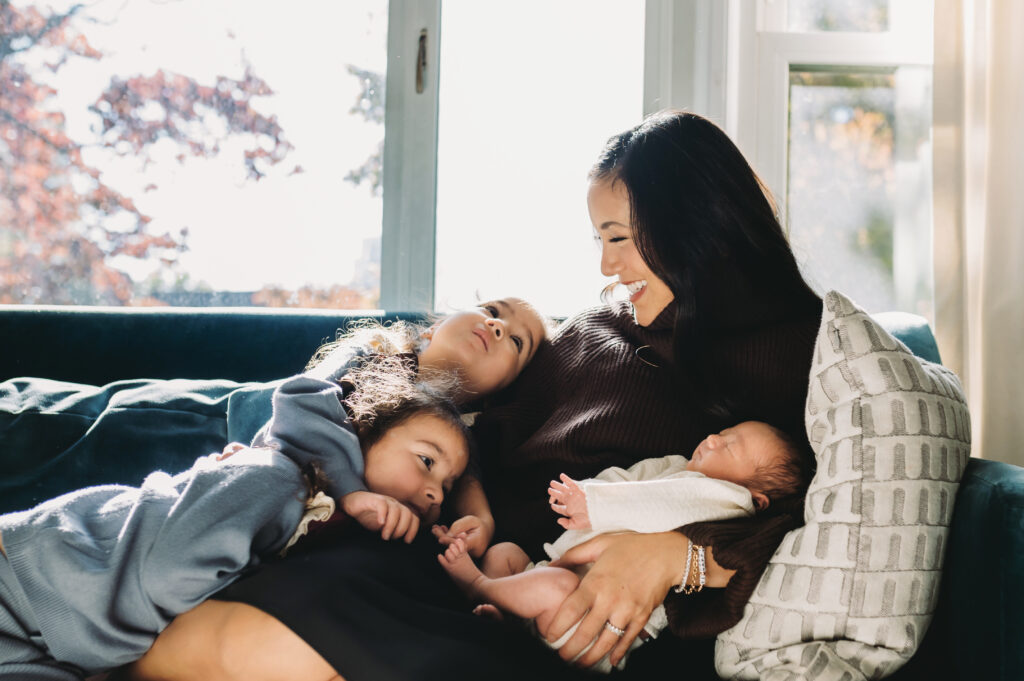 Motherhood Photography at Home: Capturing the Beautiful Chaos of Everyday Life - NPS Photography A cozy motherhood photography moment with a mom cuddling on the couch with her newborn and two older children in a sunlit living room.