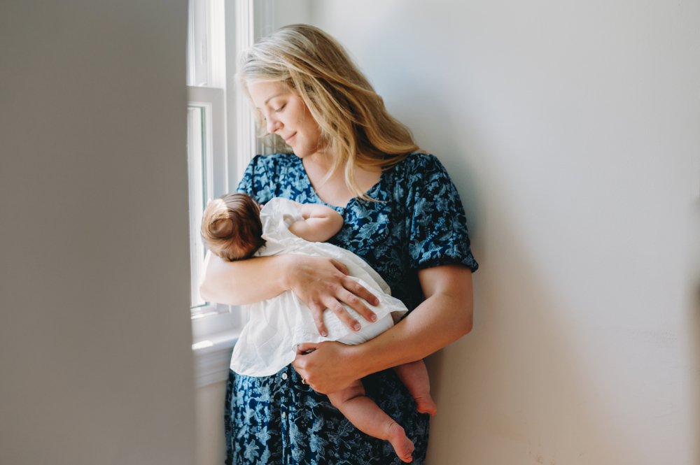 Motherhood Photography at Home: Capturing the Beautiful Chaos of Everyday Life - NPS Photography A soft moment captured during a motherhood photography session, as a mother stands by a window gently rocking her baby in natural light.