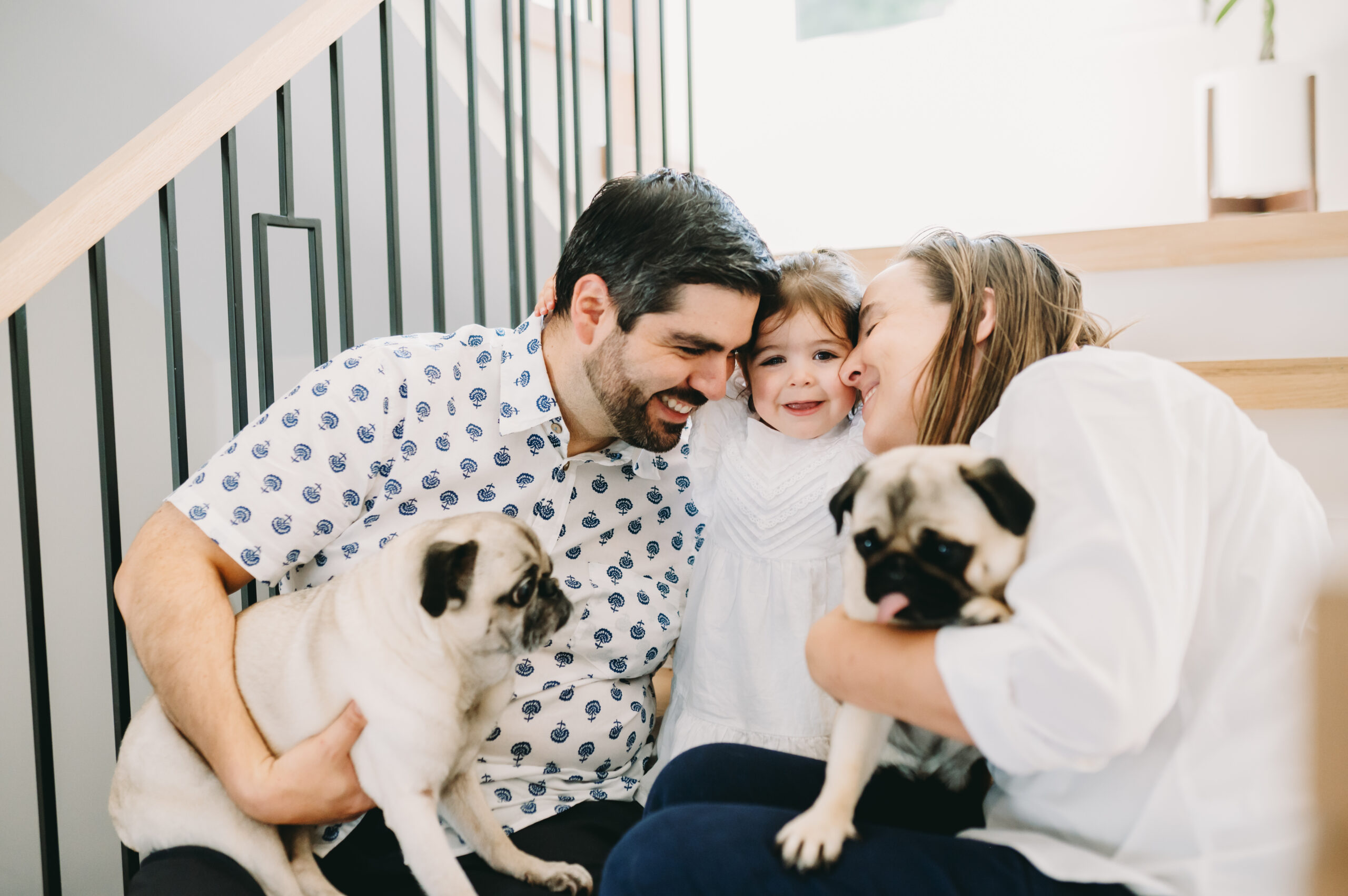 Parents and toddler smiling together with their two pugs during an in-home family photography session on the stairs. - fall photography tips