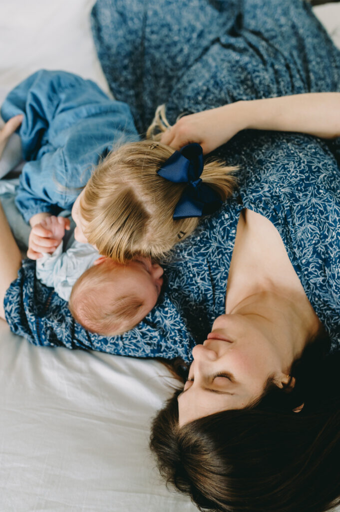 Your Top Questions About In-Home Family Photography, Answered! - NPS Photography Mother resting on bed with her newborn and toddler during an in-home family photography session capturing tender sibling love.