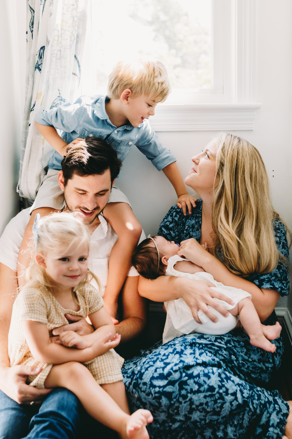 Family of five cuddling by a sunlit window during an in-home family photography session, celebrating real life and love at home.