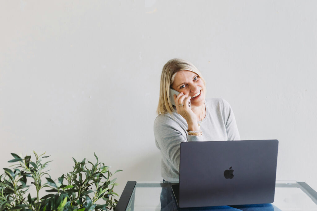 Photographer chatting on the phone while working at her laptop, demonstrating how to simplify your photography business with streamlined communication tools.