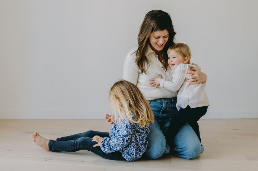 Indoor Winter Family Photos: A Cozy Home Prep Guide - NPS Photography Mother sitting on the floor hugging her toddler while her older child plays nearby, captured in a cozy, minimal studio setting for indoor winter family photos.