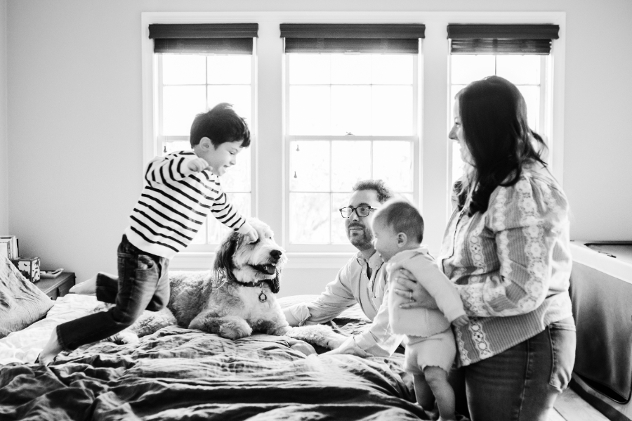 Indoor Winter Family Photos: A Cozy Home Prep Guide - NPS Photography Playful black and white indoor winter family photo of parents, two children, and their dog gathered on a bed in natural window light, showcasing candid connection at home.
