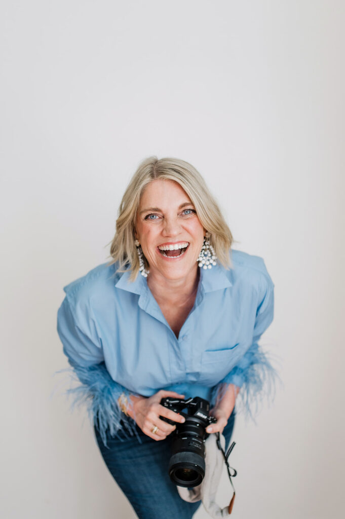 A smiling photographer holding her camera, offering a joyful look behind the lens as she connects warmly with her clients.