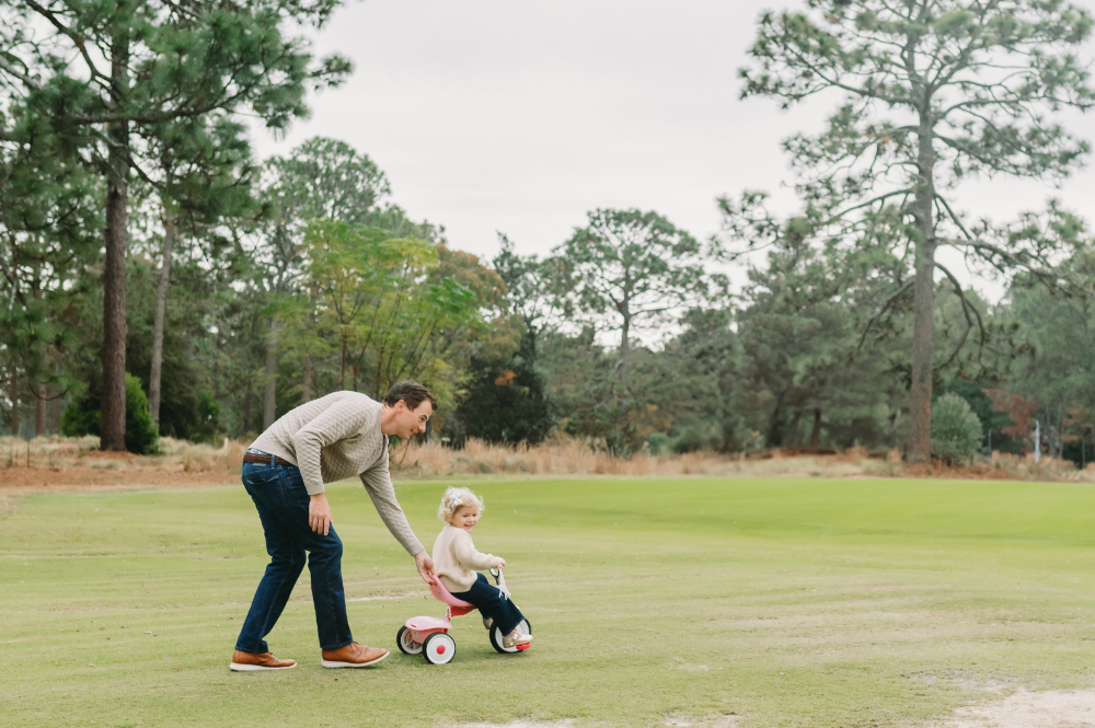 What Is Storytelling Photography? The Heartfelt Secret to Capturing Your Family’s Most Meaningful Moments - NPS Photography A dad pushing his toddler on a tricycle across an open field, capturing the beauty of outdoor freedom and the bond that grows in everyday play and illustrating what is storytelling photography on the move.